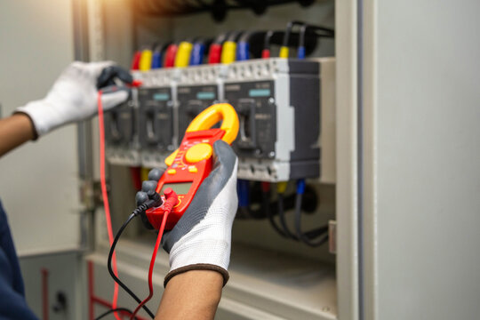 professional industrial electrician hands holding digital clamp meter and configuring test probes inside commercial circuit breaker panelboard.