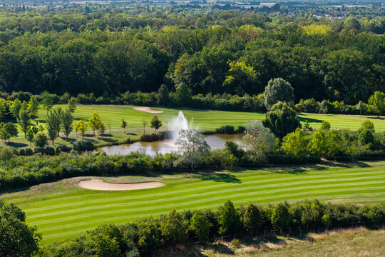 Aerial view of Magdeburg golf course shows striped fairways, sand bunkers, pond and fountain framed by dense woodland. Summer landscape combines sport, water, manicured lawns and lush greenery
