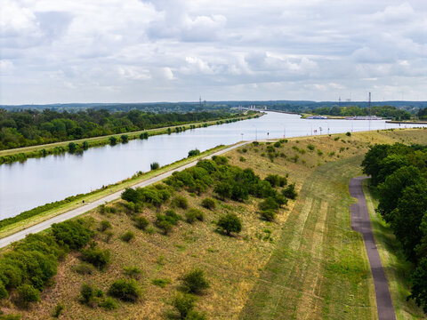 Aerial view of Mittellandkanal fork in Magdeburg with calm waterway, embankments, paths and dry summer grass. Broad canal landscape stretches beneath layered clouds, greenery and distant ships