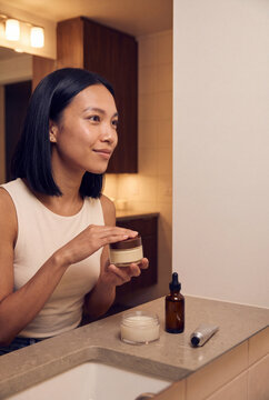 Woman applying facial cream at bathroom counter mirror for nightly skincare routine and beauty product self care banner