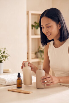 Woman arranging neutral skincare bottles on bathroom counter with towels and plants for lifestyle background and product display