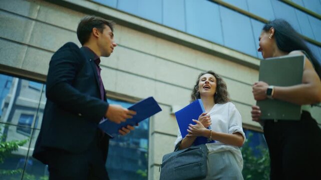 Three corporate individuals engage in an outdoor business meeting in front of a modern glass building. These colleagues hold a collaborative discussion, sharing ideas in an urban environment.