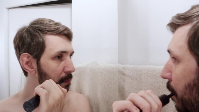 Man brushing his teeth from the side, holding an electric toothbrush near the bathroom mirror