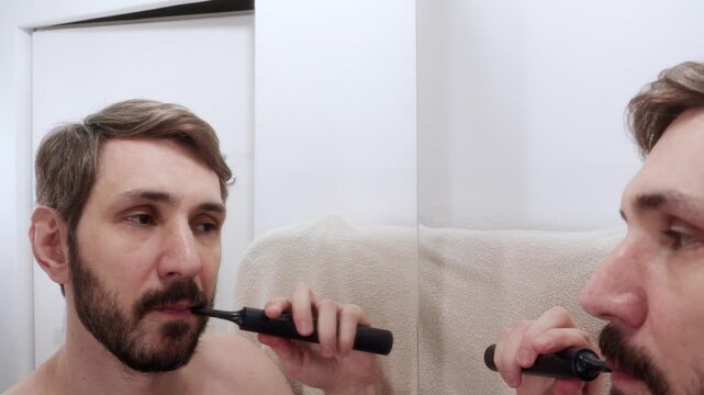 Man cleaning his teeth with a black electric toothbrush while looking into the bathroom mirror