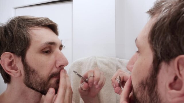 Man trimming his mustache with small scissors, looking at his reflection in the bathroom mirror