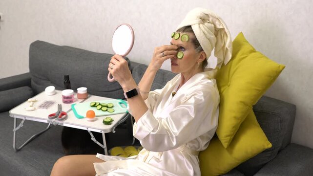 An elderly woman with a towel wrapped around her head sits on a gray couch in a white robe, applying homemade cucumber slices to her face. A small table with beauty products is beside her.