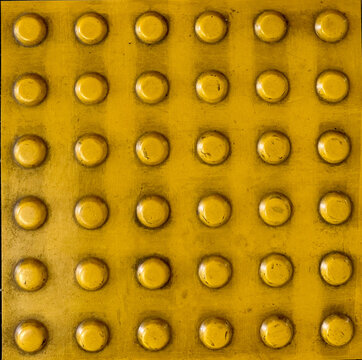 Close-up of yellow tactile paving with raised dots, showing a worn textured surface used to assist visually impaired pedestrians.

