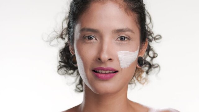 Close-up shot of black-haired hispanic beauty woman applying white clay mask on her cheek with wide brush against white backdrop | Abstract face mask and skin care application footage