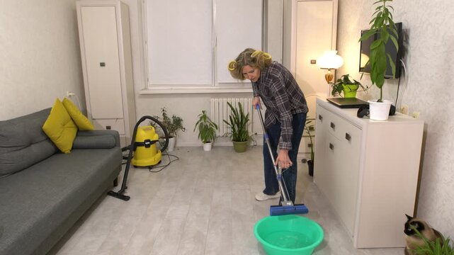 Elderly woman in plaid shirt wringing mop into green basin in tidy living room