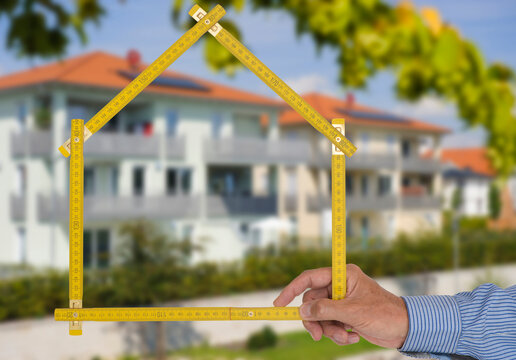 Ein Meterstab ist gefaltet in Form von einem Haus vor einem Neubau - FOTOMONTAGE