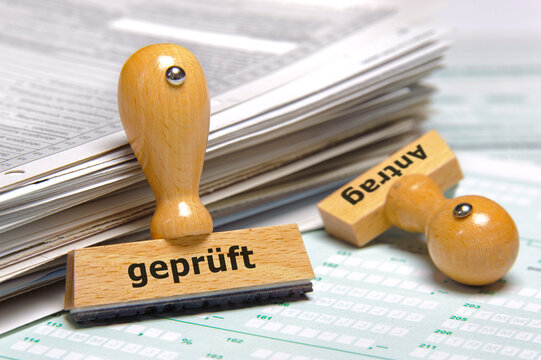 Neben einem Stapel Papiere lehnen zwei Holzstempel mit der Aufschrift: Antrag gepr&uuml;ft
