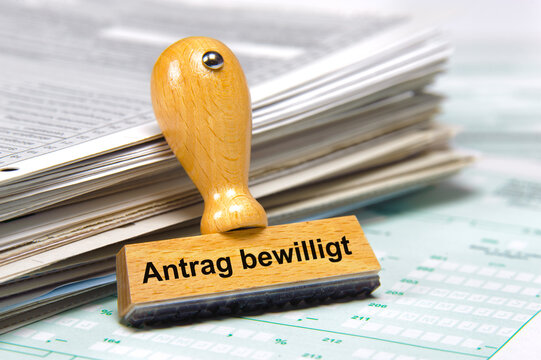 In einem B&uuml;ro lehnt ein Holzstempel an einem Stapel Papiere mit der Aufschrift: Antrag bewilligt