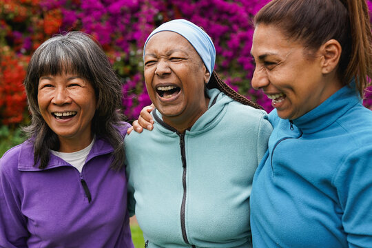 Senior women with different ethnic hugging each other at city park while laughing in front of camera - Elderly multiracial female friends having fun together outdoor