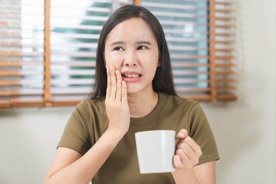 Asian young woman holding a mug and touching her cheek in pain, suffering from sensitive teeth or toothache problem while drinking hot coffee at home, dental healthcare concep.