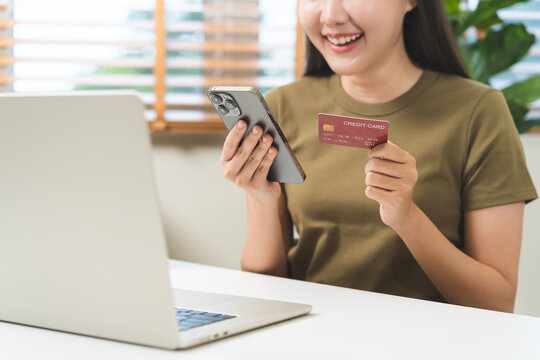 Happy asian young woman holding credit card and smartphone for online shopping payment while sitting with laptop computer at home, modern digital lifestyle and ecommerce transaction concept.