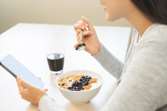 Asian young woman holding a smartphone while eating a healthy Greek yogurt bowl topped with blueberries and granola, with a cup of black coffee on the table at home.