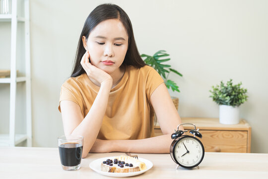 Hungry Asian woman sitting and waiting to end intermittent fasting looking bored at healthy breakfast food and alarm clock, time restricted eating nutrition plan and weight loss control concept.
