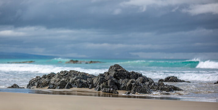 Sandstrand am Kaka Point Beach mit bewaldeter Klippe, Treibholz und Brandung unter Wolkenhimmel, Otago, Neuseeland