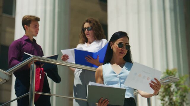 Three young business individuals stand outside a grand building, attentively reviewing documents and discussing important matters.