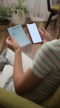 Woman gets paper prescription, sitiing on her home sofa and using smartphone to check the name of the medication online. Blank screen for design