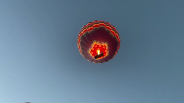 A colorful hot air balloon soars high across the clear blue sky. The burner ignites, blasting flames to heat the air inside the balloon and lifting it slowly and serenely into the heavens.