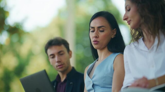 A group of young professionals holds an outdoor business meeting. They are collaborating and discussing ideas with a laptop in a park setting.