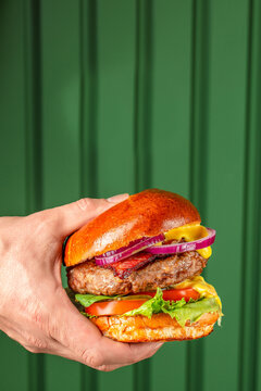 Hand holding a gourmet hamburger with lettuce, tomato, onion, and mustard against a green wooden background, showcasing a delicious meal presentation