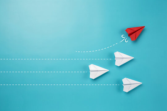 A red paper airplane breaking away from white paper airplanes on a blue background with leadership innovation strategy business direction creative different management competition development