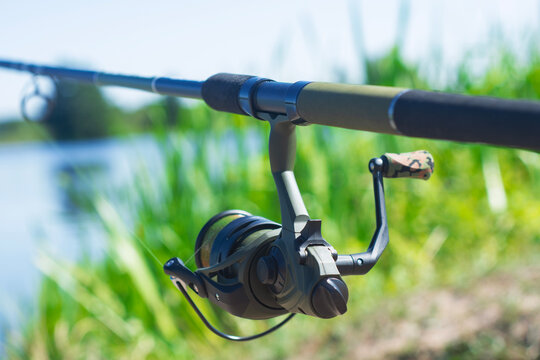 Feeder fishing rod with spinning reel near riverbank and green reeds. Fishing tackle concept for angling, sport fishing, outdoor leisure, bait feeder setup and recreational equipment advertising.