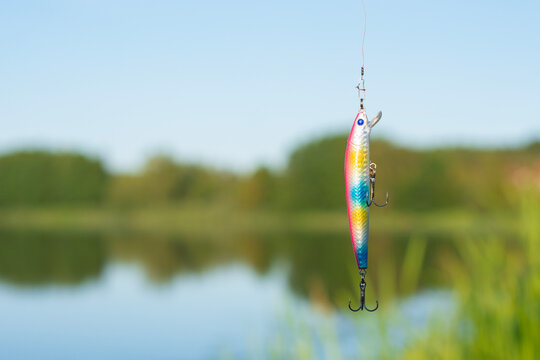 Fishing lure and colorful wobbler hanging over lake water with natural landscape background. Outdoor fishing equipment concept for angling brands and sports marketing.
