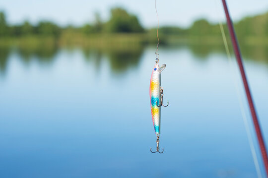 Colorful fishing lure hanging on line over calm lake with blurred shoreline. Ideal for sport fishing, lure selection, bait advertising, outdoor leisure and angling equipment promotions.