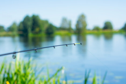 Fishing feeder rod tip over calm lake water with blurred natural landscape background. Outdoor angling scene for fishing brands, recreation marketing, sports equipment advertising and lifestyle.