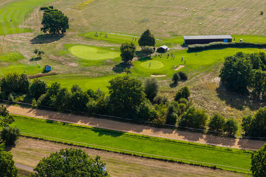 Aerial view shows golfers practicing on green course beside racetrack, fairways, bunkers and dry summer grass. Scenic recreation landscape combines sport, trees, turf and open rural countryside