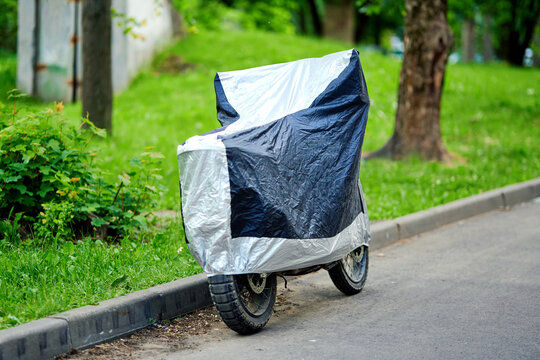 Motorcycle covered with protective tarp parked on paved road in residential area with trees and grass under natural daylight. Covered bike on street side, outdoor motorcycle storage on city street