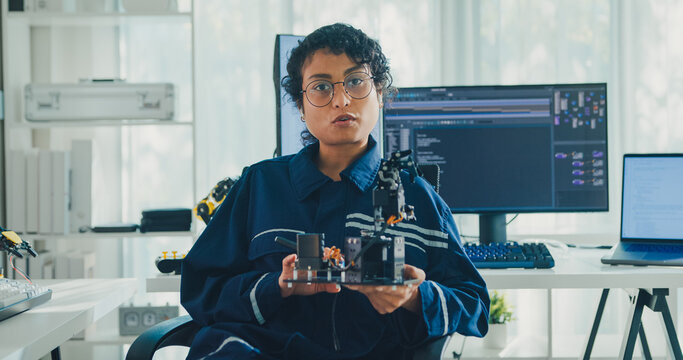 Young African American female robotics engineer discussing mechanical part on video call and looking at camera in laboratory. Remote work, virtual training, online meeting, future technology concept.
