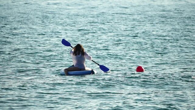 Paddleboarding woman rowing sits on her board while navigating across calm blue sea waters during a sunny summer vacation