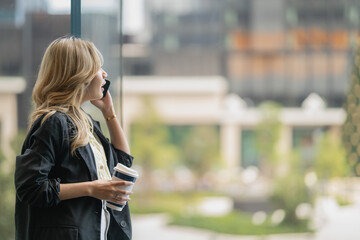 Fototapeta premium Asian businesswoman with blonde wavy hair is seen in profile talking on her phone, hair partly covering her face, holding coffee near a garden courtyard, amid the city's glass skyline.