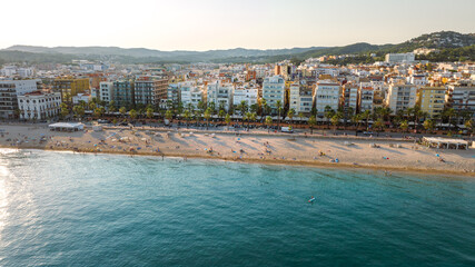 Obraz premium Aerial view of Lloret de Mar beach on Spain's Costa Brava, with sandy shore, Mediterranean Sea, palm-lined promenade, hotels and urban buildings in warm evening light.