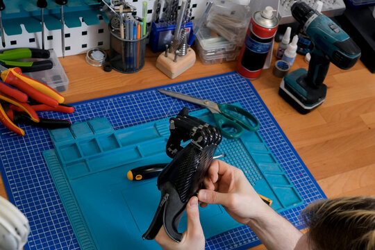 A person holds an advanced black prosthetic hand. The scene takes place on a workbench, with tools, and a blue mat in the background