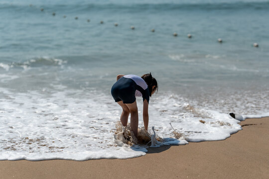 Girl bends down on a sunny beach to pick up something in ocean foam.
