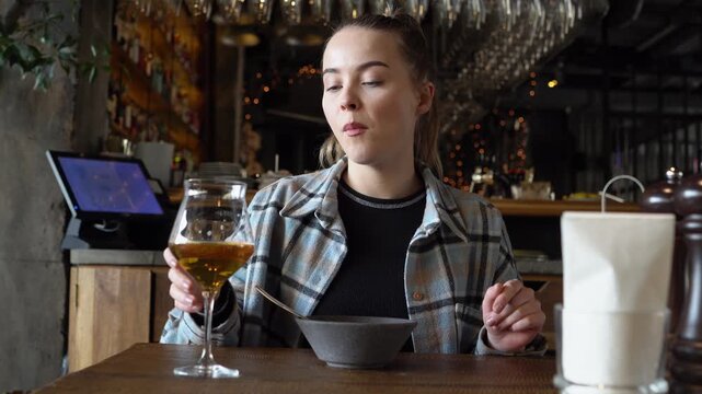 young girl eats in restaurant and drinks beer from glass, happy woman has lunch in cafe and drinks alcohol at the table