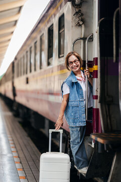 Asian older woman joyful smile travels train with luggage at bustling railway station platform ready for exciting journey public transport adventure tourism