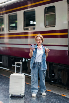 Asian adult woman traveler happily uses smartphone for digital connection preparing train journey railway station platform urban transportation adventure