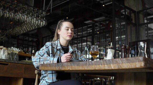 young girl eats in restaurant and drinks beer from glass, happy woman has lunch in cafe and drinks alcohol at the table