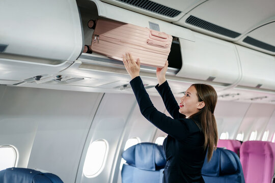 female passenger traveler places pink suitcase into overhead luggage compartment on airplane cabin during boarding travel flight journey