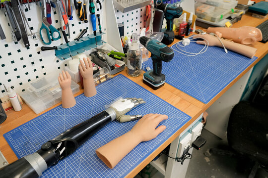 Tools hang above the work surface used to create and customize prosthetics. Various artificial hands and a partial arm rest on a gridded workbench in a well lit workshop