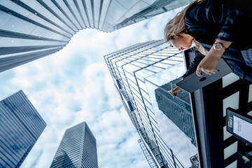 Fototapeta premium Low angle Bangkok shot of an Asian businesswoman with her head tilted forward, stylus resting on the tablet, glass towers framing a bright cloudy sky, in the afternoon light.