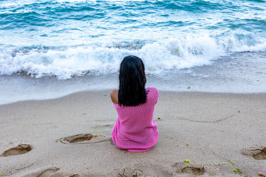 Rear view of a girl in a pink sweater sitting on sandy shore, facing gentle ocean waves, evoking calmness and reflection.