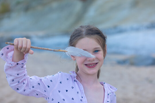 Smiling girl holding a feather on a sandy beach with soft natural light and a blurred seaside background.