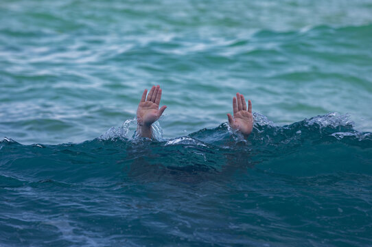 Two hands emerge from blue ocean waves, conveying urgency and emotion against a tranquil seascape, ideal for concepts of help, rescue, or struggle.